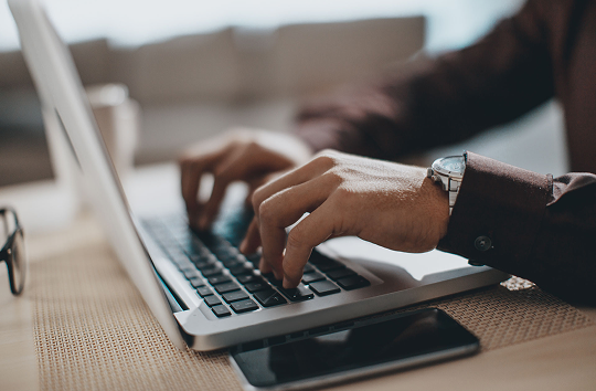 A close-up of a person's hands wearing a watch, typing on a laptop keyboard to access their HUB24 account, with a smartphone resting on the desk nearby.
