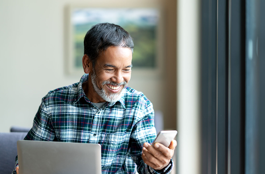 An older man with a grey beard smiling happily as he looks at his smartphone, sitting indoors near a window with a laptop open in front of him.