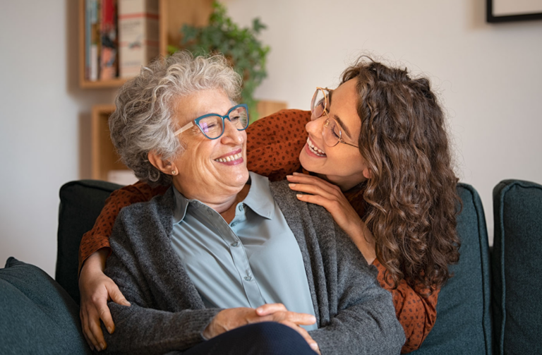 An older woman with glasses sitting on a couch, smiling warmly as her adult daughter embraces her from behind, representing the peace of mind of a HUB24 Lifetime super account.