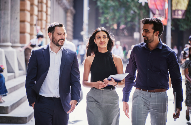 Three professionals in business attire smiling and talking as they walk together down a sunny city street, with the woman in the center holding a folder.